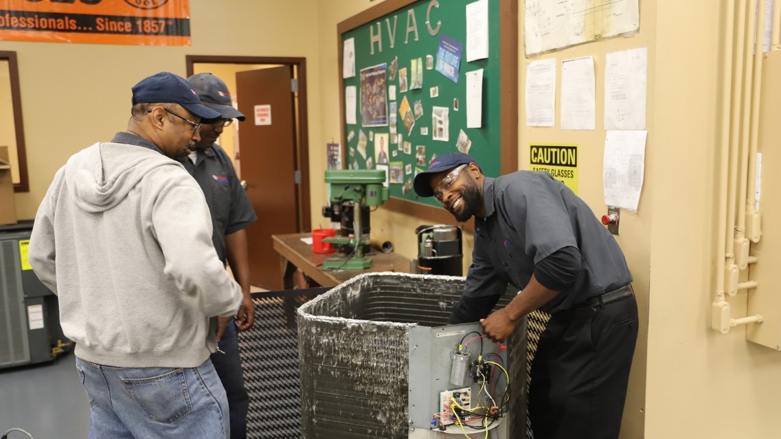 Instructor reaching into an HVAC system, students watching nearby