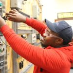 Electrical student in a red hoodie, cap, and safety glasses wiring components on a practice control panel