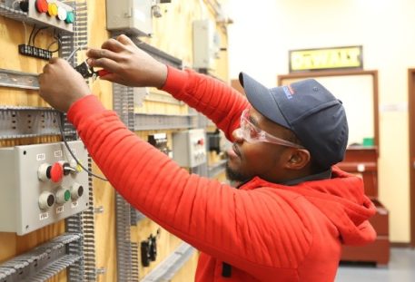 Electrical student in a red hoodie, cap, and safety glasses wiring components on a practice control panel