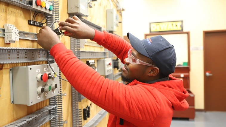 Electrical student in a red hoodie, cap, and safety glasses wiring components on a practice control panel