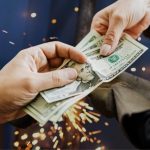 One hand passes a stack of cash to another in front of a welder at work, with sparks flying in the background.