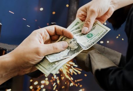 One hand passes a stack of cash to another in front of a welder at work, with sparks flying in the background.