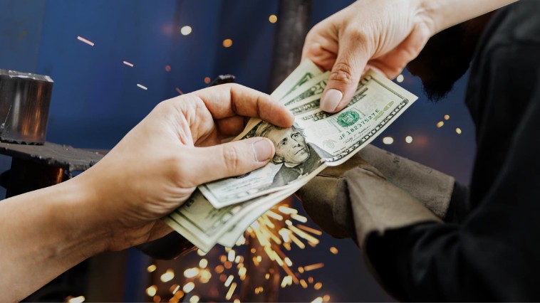 One hand passes a stack of cash to another in front of a welder at work, with sparks flying in the background.